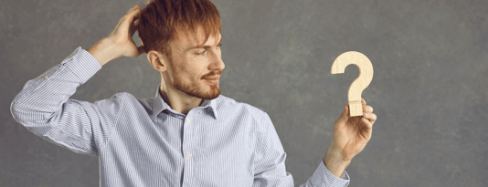 Man stands against a grey background holding onto a wooden question mark and scratching his head