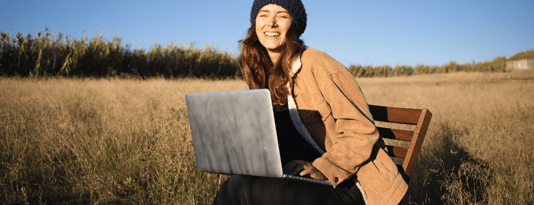 woman sits on a chair in a sunny field, smiling and working on her laptop