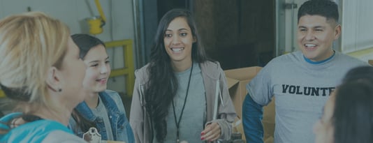 Four people engaged in conversation in what appears to be a community center or volunteer facility. From left to right: a blonde woman in a teal top, a young person in a denim jacket, a woman with long dark hair wearing a gray hoodie and lanyard, and a smiling man in a gray sweatshirt with 'VOLUNTEER' text visible. The setting has yellow structural elements in the background, suggesting an indoor community space or warehouse-style building.