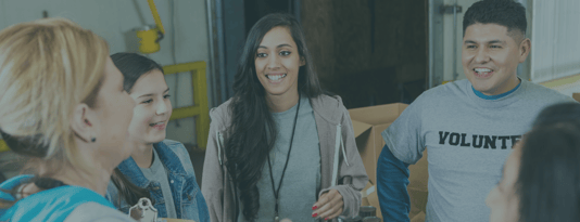 Four people engaged in conversation in what appears to be a community center or volunteer facility. From left to right: a blonde woman in a teal top, a young person in a denim jacket, a woman with long dark hair wearing a gray hoodie and lanyard, and a smiling man in a gray sweatshirt with 'VOLUNTEER' text visible. The setting has yellow structural elements in the background, suggesting an indoor community space or warehouse-style building.