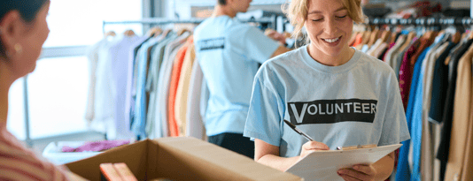 A female charity volunteer in a charity shop smiles broadly while checking an inventory on a clipboard