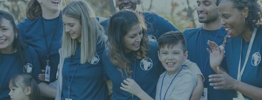 A diverse group of smiling volunteers wearing matching blue t-shirts with circular logos and blue lanyards, gathered together outdoors. The group includes adults and children of various ages and ethnicities, all appearing happy and engaged in a fundraising or volunteer event for small charity week. The lighting suggests it's during daytime with natural outdoor lighting.